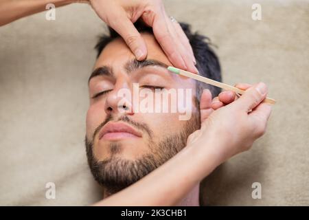 Dall'alto Crop donna usando bastone per applicare cera sul sopracciglio di giovane uomo bearded con gli occhi chiusi durante il lavoro in studio di bellezza Foto Stock