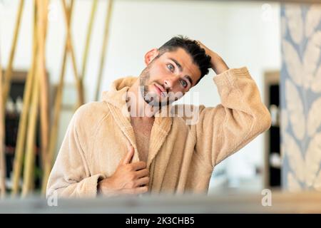 Riflesso del giovane maschio in morbido accappatoio che tocca i capelli scuri e che guarda nello specchio durante la sessione termale in salone Foto Stock