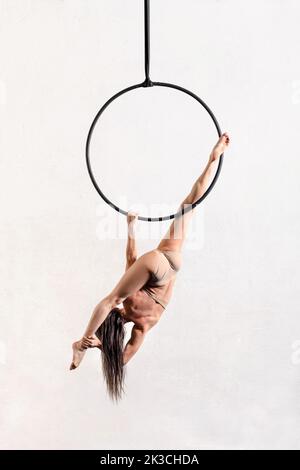 Vista posteriore del corpo intero dell'aerialista femminile anonimo che esegue la posa divisa sul cerchio dell'antenna su sfondo bianco in studio luminoso Foto Stock