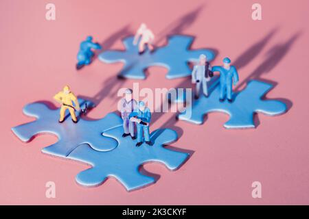 Squadra di minuscoli lavoratori in miniatura figure su jigsaw collegati puzzle pezzi isola su carta rossa, rosa. Primo piano su elementi puzzle blu collegati Foto Stock