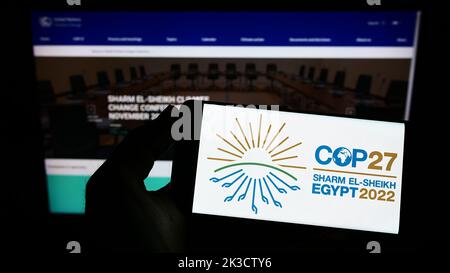 Persona in possesso di uno smartphone con il logo della 2022 Conferenza ONU sul cambiamento climatico (COP27) sullo schermo di fronte al sito Web. Messa a fuoco sul display del telefono. Foto Stock