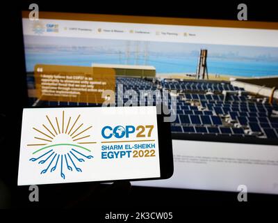 Persona che tiene il cellulare con il logo della 2022 Conferenza ONU sul cambiamento climatico (COP27) sullo schermo di fronte alla pagina web. Messa a fuoco sul display del telefono. Foto Stock