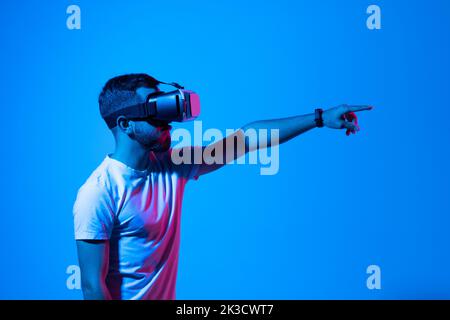 Giovane uomo barbuto che gioca a videogiochi in occhiali VR. Appassionata persona del millennio che sperimenta la realtà virtuale mentre si trova in piedi nella luce al neon, copia spazio. Foto Stock