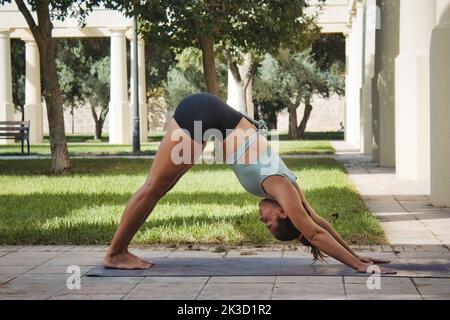 Una giovane donna che indossa pantaloncini corti e crop top che fa yoga benessere in un parco pubblico in una giornata di sole in estate su un sentiero vicino all'erba Foto Stock