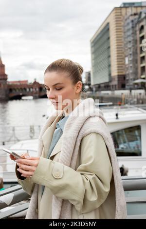 Donna in trench coat utilizzando il telefono cellulare su strada urbana a Berlino, immagine stock Foto Stock