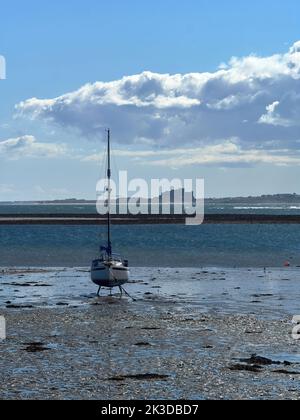 Uno yacht arroccato sulla sua chiglia nella zona intertidale, con il Mare del Nord e il Castello di Bamburgh dietro e un elaborato paesaggio nuvoloso sopra. Foto Stock