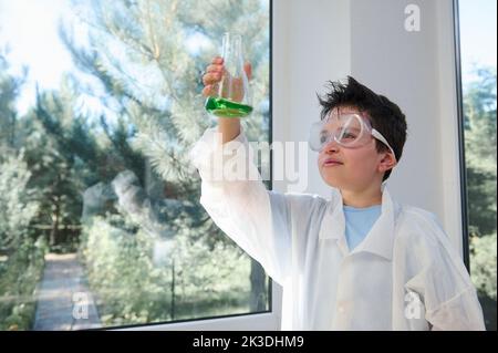 Adorabile bambino con capelli scuri che indossa occhiali di sicurezza e camice da laboratorio per esaminare la reazione chimica in corso nel pallone Foto Stock