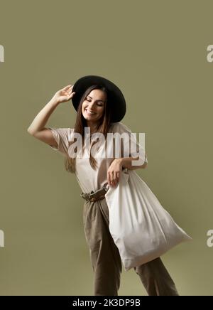 Donna sorridente in una t-shirt verde e cappello nero sta tenendo il tessuto di tela del sacchetto isolato su fondo verde. Foto Stock