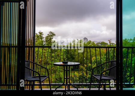 Vista dalla finestra dell'hotel con balcone sulla foresta tropicale durante la tempesta con la pioggia. Grandi nuvole d'ombra e tempo piovoso. Due sedie sul lato wi Foto Stock