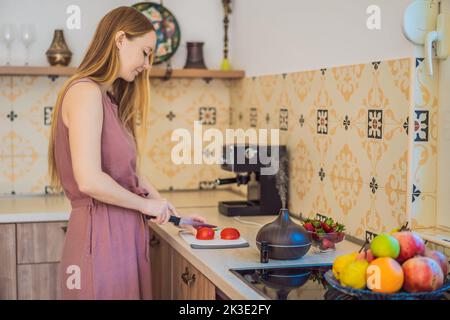 La donna prepara il cibo accanto a un diffusore di aroma. La cuoca taglia i peperoni rossi maturi in cucina. Cucina insalata di peperoni freschi, maturi per gli ospiti. Estate Foto Stock