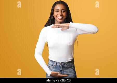 Sorridente giovane afroamericana con capelli afro indossare casual elegante su sfondo giallo tenendo vuoto con le mani, misurare simbolo, sorridere e guardare la macchina fotografica, pubblicità concetto Foto Stock