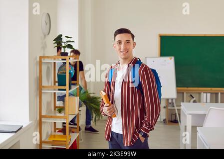 Ragazzo studente in camicia a righe con zaino e libro in classe sorridendo e guardando la macchina fotografica. Foto Stock