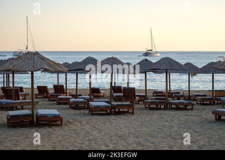 IOS, Grecia - 11 settembre 2022 : lettini, ombrelloni e barche a vela sulla spiaggia di Mylopotas mentre il sole tramonta lentamente Foto Stock