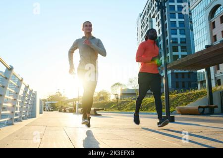Giovane uomo e donna interculturale in giacche sportive, leggini neri e sneakers jogging lungo il ponte e lungo il fiume al mattino Foto Stock
