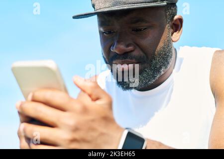 Primo piano del volto di giovane sportivo afro-americano bearded in t-shirt bianca e berretto da baseball nero che scorre nel telefono cellulare all'aperto Foto Stock