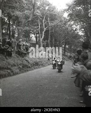 1954, storico, in una sezione boscosa di una foresta, che si affaccia su una strada sul circuito di Mount Oliver, gli spettatori in attesa applaudono due motociclisti vittoriosi, con rossette di fiori su di loro, al termine delle gare di Scarborough, Inghilterra, Regno Unito. Il famoso circuito di Oliver's Mount è l'unico circuito naturale dell'Inghilterra, conosciuto come "mini TT". L'impegnativo tracciato ondulato si trova vicino alla spiaggia di Scarborough, da cui le gare erano tradizionalmente conosciute come le Scarborough Races. Foto Stock