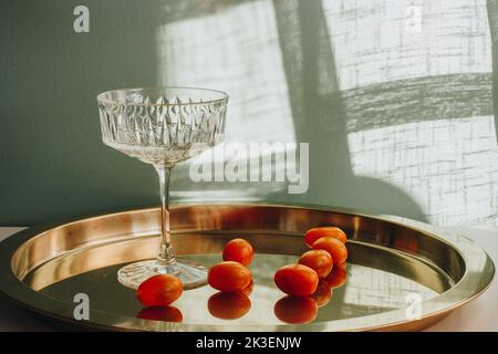 un bel vetro di cristallo con pomodori rossi si erge su un vassoio di metallo dorato illuminato dalla luce del sole, gettando un'ombra su una parete bianca. Foto Stock