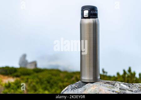 Thermos di acciaio su una pietra. Bottiglia di acciaio su una pietra in montagna. Bottiglia sullo sfondo di nuvole e montagne. Vista frontale. Foto Stock