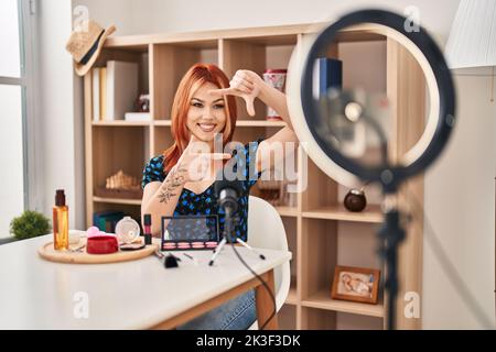 Giovane donna caucasica applicare make up fare tutorial sorridendo rendendo cornice con mani e dita con il volto felice. Creatività e fotografia concetto. Foto Stock