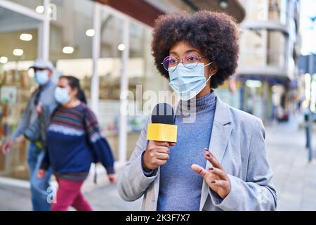 Donna afroamericana reporter indossare maschera medica utilizzando il microfono che lavora per strada Foto Stock