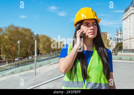 giovane donna venezuelana caucasica latina, ingegnere con casco giallo, giubbotto e abiti blu, all'aperto con dubbio parlando con il suo capo al telefono, tecnologia Foto Stock