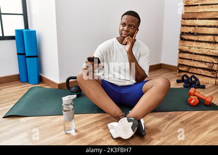 Giovane uomo africano seduto sul tappeto di allenamento in palestra con smartphone con mano sul mento pensando alla domanda, all'espressione penitente. Sorridendo con te Foto Stock