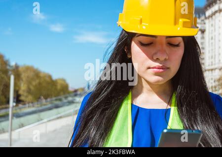 giovane donna ingegnere venezuelano caucasica latina con casco giallo, giubbotto e abiti blu, all'aperto leggere un messaggio di testo con istruzioni da lei Foto Stock