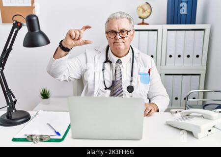 Uomo caucasico anziano che indossa uniforme medico e stetoscopio in clinica sorridente e sicuro gesturing con mano facendo segno di piccole dimensioni con le dita Foto Stock