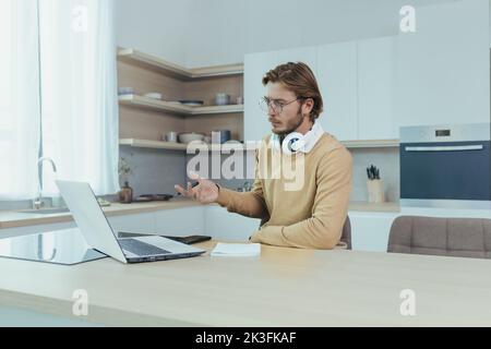 Giovane uomo d'affari che lavora a distanza da casa seduto in cucina, uomo che utilizza il computer portatile per videochiamare concentrandosi e parlando seriamente in una riunione online con i colleghi colleghi. Foto Stock