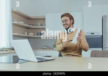 Un insegnante giovane insegna a distanza da casa, l'uomo lavora in cucina a casa, usa il laptop per le chiamate online e comunica con gli studenti sorridendo spiega l'attività mostra il notebook. Foto Stock