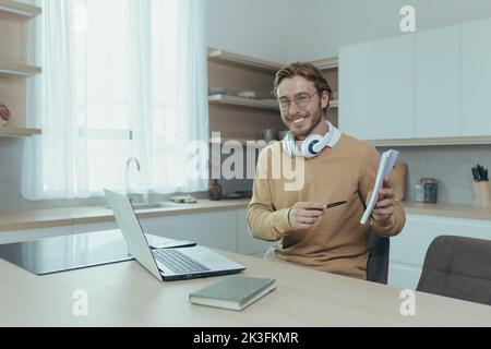 Un insegnante giovane insegna a distanza da casa, l'uomo lavora in cucina a casa, usa il laptop per le chiamate online e comunica con gli studenti sorridendo spiega l'attività mostra il notebook. Foto Stock