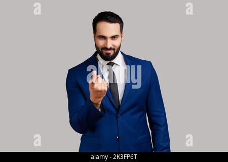 Ritratto di positivo sorridente bell'uomo d'affari bearded che chiama con un gesto delle dita, invitando a entrare, indossando tuta ufficiale. Studio in interni isolato su sfondo grigio. Foto Stock