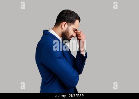 Vista laterale di triste sconvolto infelice uomo d'affari bearded che nasconde faccia in mano piangendo, sentendosi sollecitato preoccupato, esprimendo dolore, indossando il vestito ufficiale di stile. Studio in interni isolato su sfondo grigio. Foto Stock