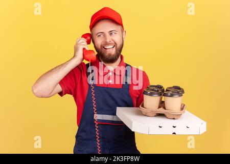 Ritratto di uomo corriere in tuta t-shirt rossa e cappello parlare telefono fisso e caffè con pizza, chiamata per ordinare fast food. Studio al coperto isolato su sfondo giallo. Foto Stock