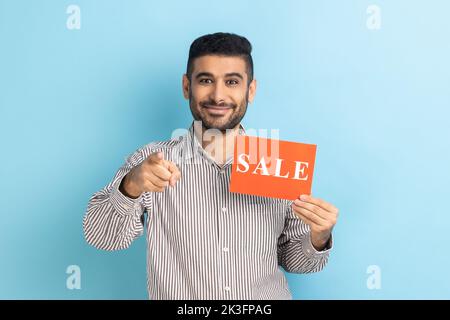 Ritratto di uomo d'affari soddisfatto sorridente che indica la macchina fotografica e la scheda di tenuta con l'iscrizione di vendita, prezzi bassi sul venerdì nero, che indossa la camicia a strisce. Studio in interni isolato su sfondo blu. Foto Stock