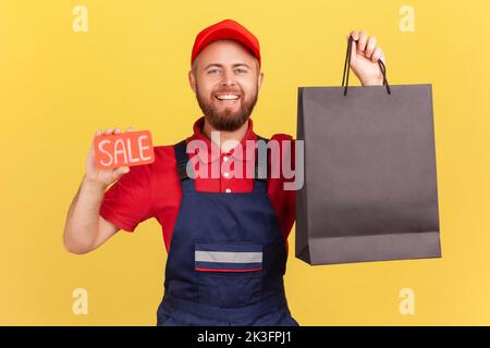 Ritratto di felice felicissimo corriere uomo guardando la macchina fotografica con un sorriso toothy, tenendo la borsa nera della spesa e la parola di vendita, indossando tute blu. Studio al coperto isolato su sfondo giallo. Foto Stock