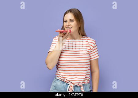 Ritratto di una donna bionda positiva sorridente e felice che indossa una T-shirt a righe in piedi con smartphone e registrazione di messaggi vocali, assistente vocale. Studio al coperto isolato su sfondo viola. Foto Stock