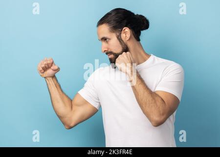Vista laterale di un uomo aggressivo con barba che indossa una T-shirt bianca con pugni aggrappati pronti a pugilato, allenatore di arti marziali, autodifesa. Studio in interni isolato su sfondo blu. Foto Stock