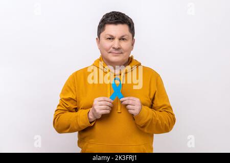 Consapevolezza del cancro alla prostata. Uomo serio che tiene il nastro blu nelle sue mani, sostiene la gente che vive e la malattia, con la felpa con cappuccio di stile urbano. Studio in interni isolato su sfondo bianco. Foto Stock