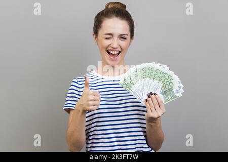 Ritratto di donna che indossa una T-shirt a righe che tiene un mazzo di banconote in euro e che strinca, mostrando un gesto di pollice in su, segno di successo, approvazione. Studio in interni isolato su sfondo grigio. Foto Stock