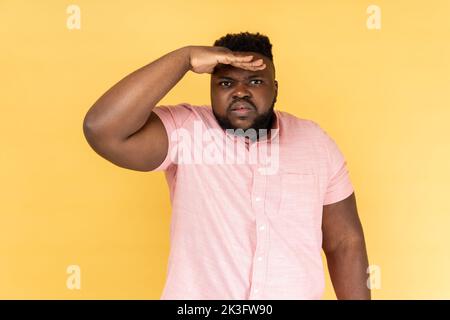Ritratto di curioso uomo con barba in camicia rosa tenendo palmo sopra la testa e guardando attentamente lontano, peering con aspettativa a lunga distanza. Studio al coperto isolato su sfondo giallo. Foto Stock