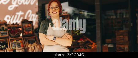 Il proprietario riuscito del supermercato sorridente alla macchina fotografica mentre sta in piedi nella sezione di prodotti freschi del suo deposito della drogheria. Felice giovane donna che gestisce una piccola Foto Stock