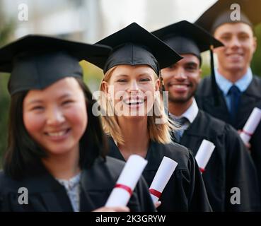 Lasci le nuove avventure cominciare... Un gruppo dei laureati che si levano in piedi nel tappo e nel abito e che tengono i loro diplomi. Foto Stock