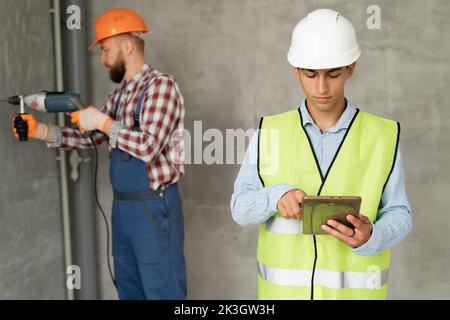 ingegnere o architetto che indossa giubbotto di sicurezza e caschi, ispezionando il progetto lavorando con un computer tablet in cantiere. Operai edili w Foto Stock