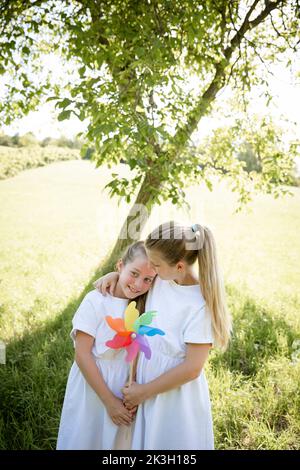 due belle ragazze, sorelle con abiti bianchi che tengono colorati ruote e in piedi in verde prato di fronte a un albero di noce e sono felici Foto Stock