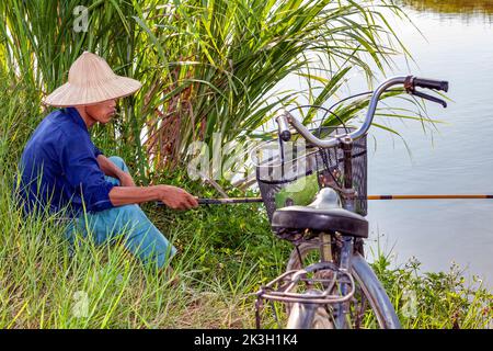 Contadino vietnamita che indossa cappello di bambù con bicicletta e canna da pesca in risaia, rurale Hai Phong, Vietnam Foto Stock
