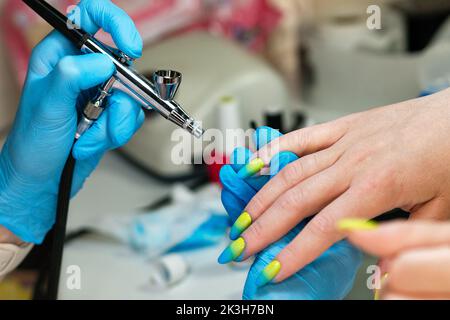 Manicurista utilizzando aerografo per dipingere unghie cliente femminile in salone unghie. Il padrone fa le unghie di disegno con aerografo. Donna cliente che ottiene manicu unghie Foto Stock