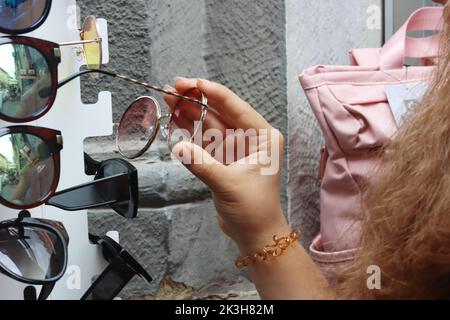 Persona che sceglie gli occhiali da sole dall'esposizione di strada nel negozio ottico. Occhiali da sole con supporto per le mani. Giovane donna che raccoglie gli occhiali da sole dalla strada stand in op Foto Stock