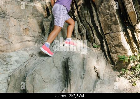 Foto delle gambe di un bambino che si arrampica sulla roccia in montagna, vista laterale. Bambina in pantaloncini sportivi e sneakers scendere ripida pendenza per percorso pericoloso Foto Stock