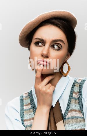 giovane donna pensiva con trucco in beretta e maglia gilet isolato su grigio, immagine stock Foto Stock